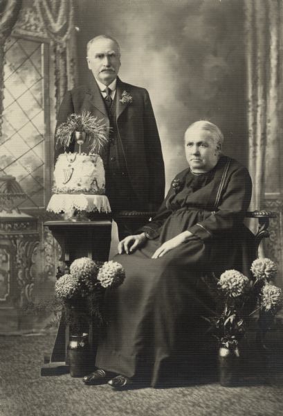 Full-length studio portrait in front of a painted backdrop of an elderly couple for their 50th anniversary. The woman is sitting, and the man is standing. Flowers are in vases on the floor, and a cake is on a small table.