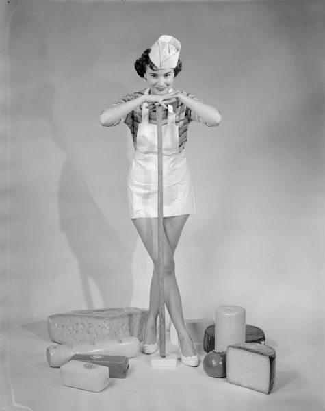 Promotional studio portrait of a woman standing and wearing a striped shirt, short apron, a paper hat and high heels, posing with her chin on her hands on top of a long stick propped on a block of wood on the floor. Various cheeses are displayed on the floor around her feet.