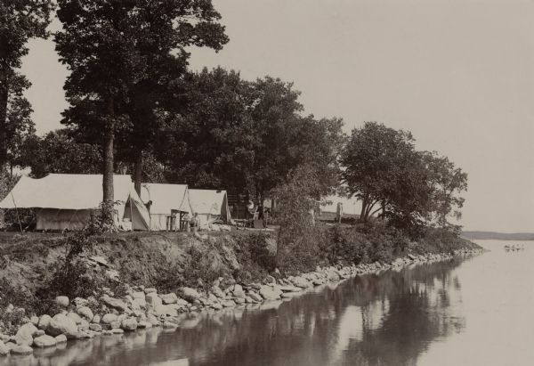 Campers on Picnic Point. Tents are set up along the shore, under the trees. A few campers are in front of their tents. A canoe is barely visible on the right. Along the shoreline is rock rip-rap.