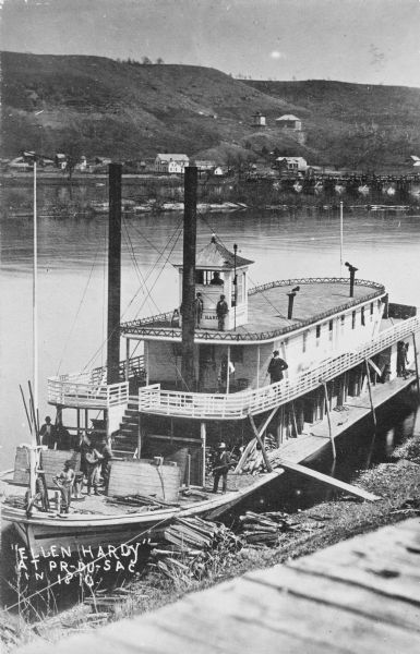 Elevated view of the steamer <i> Ellen Hardy</i> at dock. Text on the bottom left reads: "'Ellen Hardy' at Pr-Du-Sac. in 1870." The <i>Ellen Hardy</i> belonged to businessman Holmes Keyser. At least 12 men can be seen on the three decks. In the background is the river, opposite shore, Prarie Du Sac bridge and buildings below the hills. The building at upper right would later become the Wollersheim Winery.
