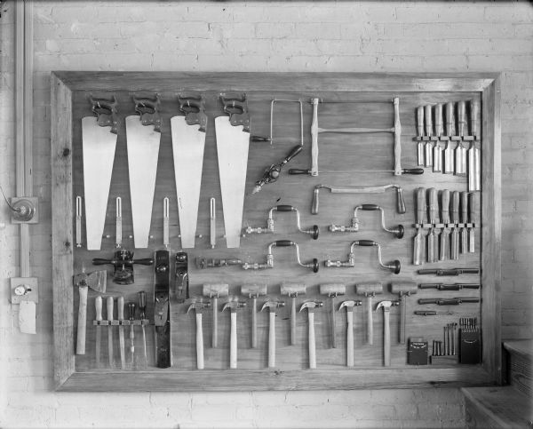 A well-organized tool board with a variety of implements: saws, drills, bits, squares, braces, chisels, scrapers, planes, wrenches, an ax, screwdrivers, mallets, awls and hammers. The tool board has a wooden frame and is hung on a brick wall. On the left are electrical outlets and on the right are wooden steps.