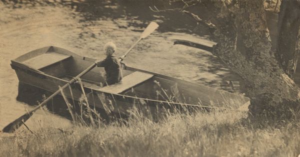 View from grassy shoreline looking down at James Peterson, as a young boy, sitting in a rowboat looking over his shoulder back towards the shoreline. He is holding one oar up and one oar down. The bow, obscured by a large tree, is probably tied up to the shore.