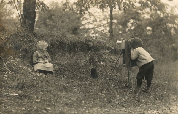 Laurie Peterson pretending to take a photograph, with a prop camera, of Muriel Peterson as she is gazing at a cat. In the background are trees and shrubs. Caption on back reads: "The Amateur."