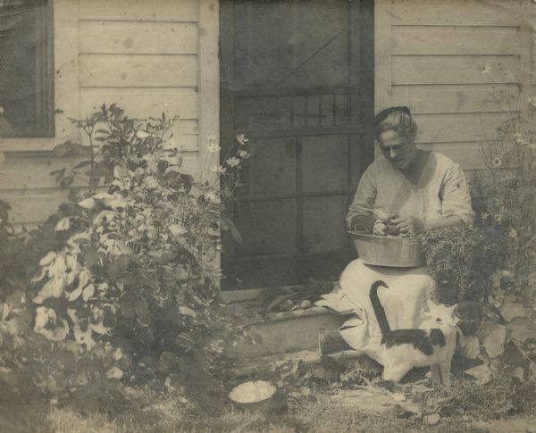 Mrs. (Christine) Peterson by the back door. She is sitting on the top step and a cat is standing at her feet. A small pile of vegetables are next to her on the step, and she has a pan on her lap. Flowers are growing on the left and right of the door. A quote from the book <i>A Life in Photographs and Letters, Carl & Christine Peterson, My Grandparents</i>, "She was always helping others. She made the coffee for church socials, and being very healthy, was often called on to take care of relatives who were sick."