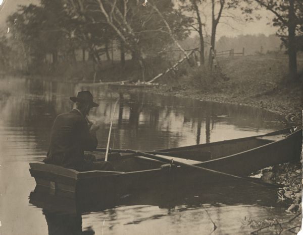 View from shoreline of the photographer, Carl A. Peterson, taking a moment to light his pipe while fishing from a boat near the family home.