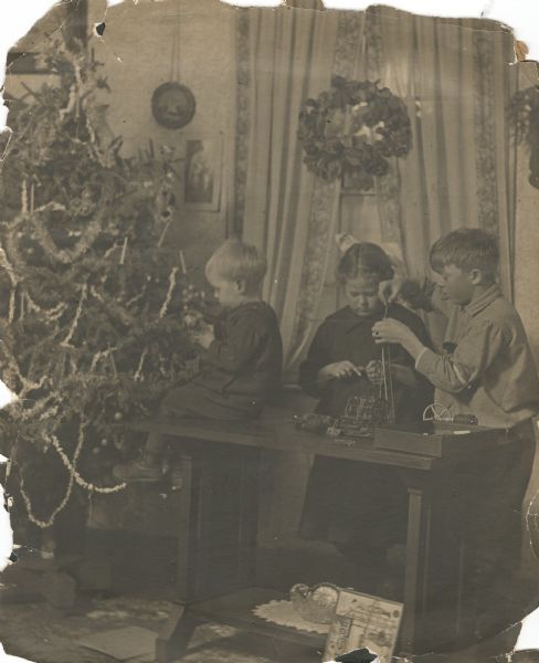 Laurie and Muriel playing with an erector set, while James is sitting on the end of the table facing the decorated Christmas tree. More toys are on the floor. A Christmas wreath hangs in the window in front of the drapes.