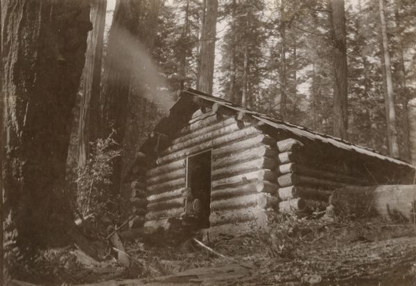 A log cabin out west in a forest. View looking up hill towards a man sitting in the open doorway with a gun in his lap. Smoke is above the cabin. At this time Carl Peterson was writing letters home to Christine Jenson about his journey. A note on the back indicates that it is Christmas.