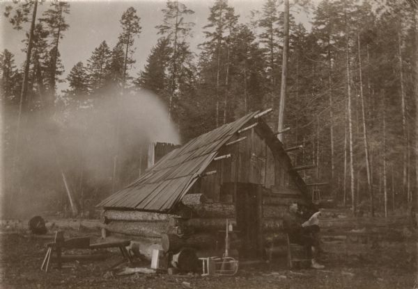 View of a man sitting outdoors in a chair near the door of a log cabin in a clearing. He is smoking a pipe and reading. A lamp is hanging from the end of a rafter, and smoke is pouring out of the chimney at the back of the cabin. On the left side of the door is a chair laying on its side near a gun, and a cutting bench, also known as a "schnitzelbank," a word of German origin. At this time Carl Peterson was writing letters home to Christine Jenson about his journey.