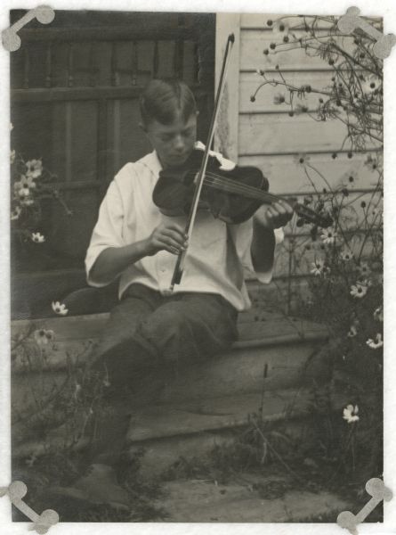 Laurie Peterson playing the violin while seated on the top step in front of a screen door. Flowers are growing on both sides of the steps.