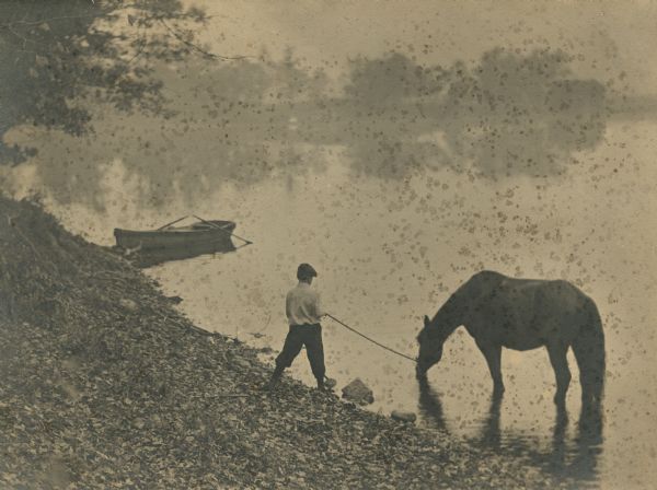 Laurie Peterson watering his horse in the lake. The sloped shoreline is covered with rocks and leaves. Farther up the shoreline is a rowboat with its oars in the water. Fog, trees and their reflections are in the background. Caption on back reads: "His Morning Drink."