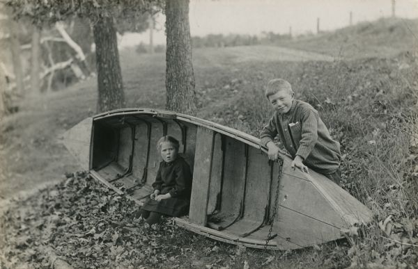 Laurie and Muriel Peterson posing with a wooden rowboat that has been tipped on its side. Laurie is standing behind the boat, leaning on one arm. Muriel is sitting inside the boat.