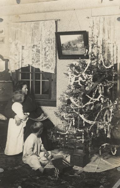 Christine, Laurie and Muriel Peterson with their Christmas tree. Christine (mother) is sitting in a chair with her arm around Muriel who is standing next to her, and Laurie is sitting on the floor. A few toys are under the tree, and a framed painting hangs on the wall between two windows with lace curtains. Caption reads: "Waiting for Santa Claus."
