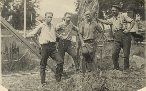 Group portrait of four telephone linemen standing together, three of them wearing pole climbing spikes (or spurs). The man leaning on the pole is Carl Peterson, the first man on the left is Laurie Peterson, son of Carl. On the right in the background is a truck. A shovel is blade down in a dirt pile in the foreground. The men worked for the Scandinavia-Iola Telephone Company.
