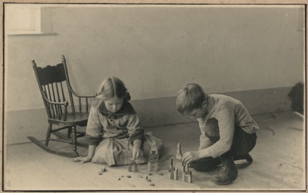 Laurie and Muriel Peterson playing with blocks on the floor. Behind Muriel is a rocking chair below a window. Handwritten on the reverse: "Interested."