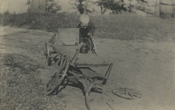 James Peterson, as a young boy, fixing a dilapidated wagon outdoors. He is attempting to reattach the wheel. A can of grease is sitting on the wagon bed.