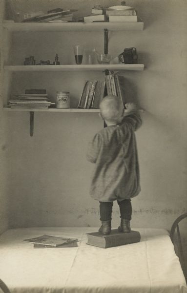 James Peterson, as a toddler, standing on a book on a table and reaching for an object on the bottom of a set of three shelves. Items resting on the shelves; mortar and pestle, camera, liquid in a glass, books, magazines, can of paste, ink bottles, plates, and a rubber stamp and ink pad. James is the youngest child of the photographer. Handwritten on reverse: "Inquisitive."