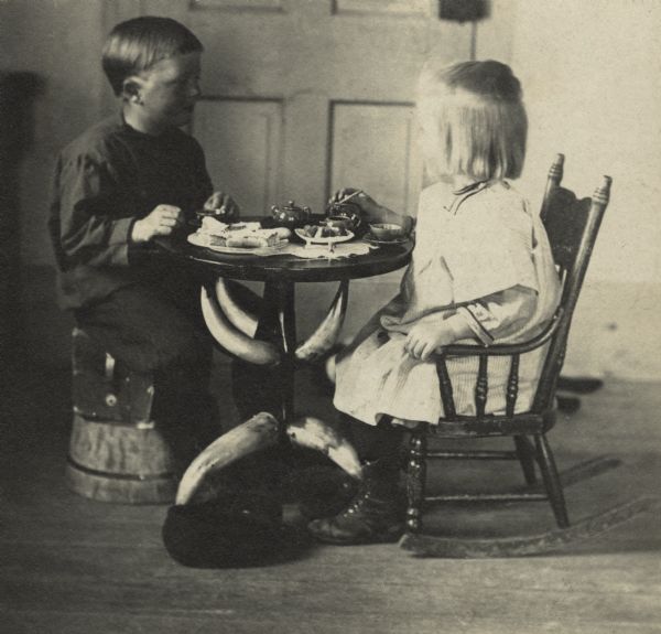 Laurie and Muriel Peterson having a tea party. In addition to tea, there are plates of pastries. Muriel is sitting in a rocking chair and Laurie is perched on a box set on an upside down tub. The table legs and supports appear to be made with cattle horns. In the background is a closed door.