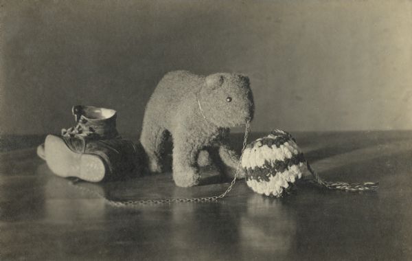 Still life of a stuffed bear, a knitted ball, and children's shoes. The stuffed bear has a ring in his nose, with a chain attached. Handwritten on reverse: "The Guardian, Jan. Still Life, 329, C.A. Peterson, Scandinavia, Wis."<p>Caption in book reads, "The photos on the next few pages include pictures of James, son of Carl and Christine, born October 31, 1912. James died October 30, 1916, one day before his fourth birthday after falling into a washtub full of boiling water when his mother was washing clothes. This tragedy was never talked about, since Christine thought it was her fault. I wasn't aware of James since there were no pictures of him at her house. These photos were only brought out after she died in 1967."