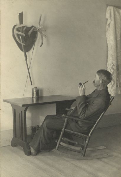 Carl Peterson relaxing in a rocking chair at his desk. He is smoking a pipe and gazing up at a pair of mounted deer horns holding a fishing pole and rifle. A canister of tobacco rests on the desk top. Underneath the desk on a low shelf is a statue, perhaps a pipe rest. Handwritten on the reverse: "Reverie, Carl A. Peterson, Scandinavia, Wis."