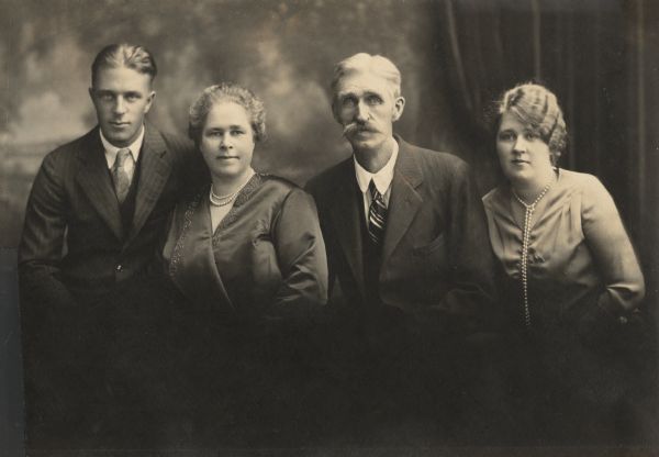 Formal waist-up group portrait of the Peterson Family. Names (l to r), Laurie Peterson, Christine Peterson, Carl Peterson, and Muriel Peterson. In the background is a painted backdrop. Laurie and Muriel left for college about this time.