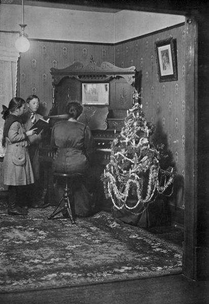 Muriel and Laurie singing as their mother, Christine Peterson, plays the organ at Christmas. On the right is a decorated Christmas tree. A large area rug covers the wood floor.