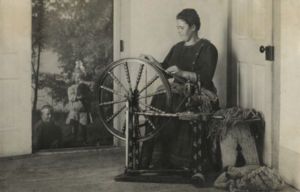 Christine Peterson working at her spinning wheel. Watching her from the open doorway are Laurie and Muriel, Christine's children. Muriel is holding a black cat.