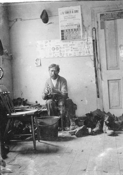 A cobbler repairing a boot in his shop. Many boots are lined up on the floor, and a bench of tools is behind him. A calendar from J.B. Yunker & Sons hangs on the back wall, along with a chart of tools and a card of a woman in a hat. Carl was always willing to take a photograph when asked.