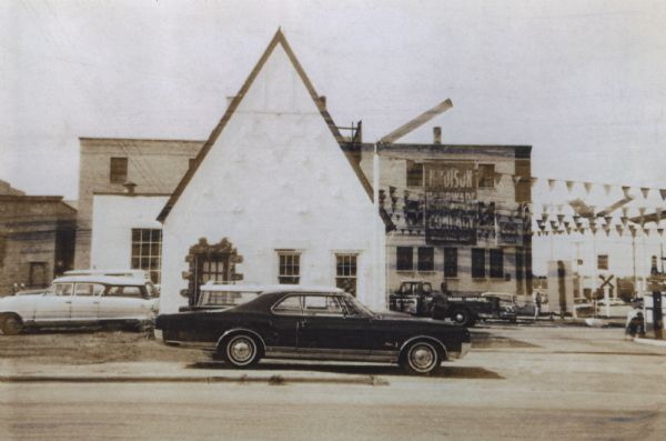 Service station located at 601 West Washington Avenue, on the intersection with South Bedford Street. This is the view from South Bedford Street. In the background is the Madison Hardware Company building. The automobile parked at the side of the building is a new 1965 Oldsmobile, and behind it is a service truck. Flag banners are hanging from the front of the station.