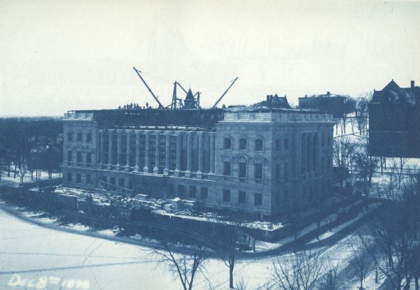 Elevated view across Langdon Street of the Wisconsin State Historical Society building under construction in winter with snow on the ground. A group of men are standing on the roof, and a crane is mounted on a open floor below them. Bascom Hill is in the background, and Science Hall is on the right.