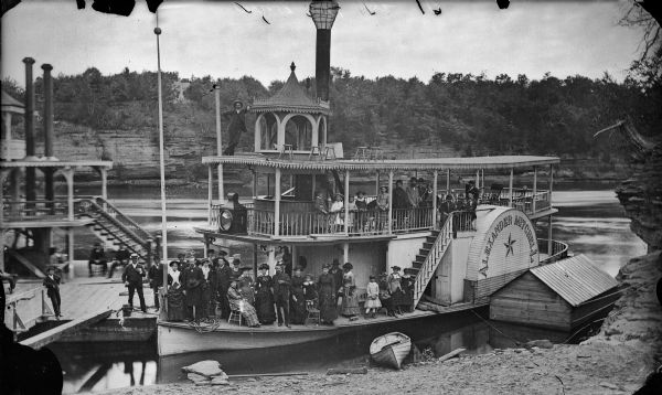 Elevated view of the steamboat <i>Alexander Mitchell</i> and the back of a second unidentified steamboat at a dock. Captain Dave and most of the passengers are posing sitting and standing. There is a rowboat on the sandy shoreline in the foreground. Bluffs and trees, and what may be a house, are on the opposite shoreline.