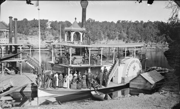 Elevated view of the steamboat <i>Alexander Mitchell</i> at a dock. Captain Dave and most of the passengers on the lower level are posing standing. There is a rowboat on the sandy shoreline in the foreground. Bluffs and trees, and what may be a house are on the opposite shoreline.