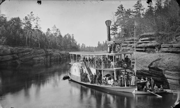 Elevated view across water towards the steamboat <i>Alexander Mitchell</i> moored in the canyon along the shoreline. Captain Dave and passengers are posing on board, with distinctive Dells rock formations in the background. In front of the steamer is a rowboat with passengers.