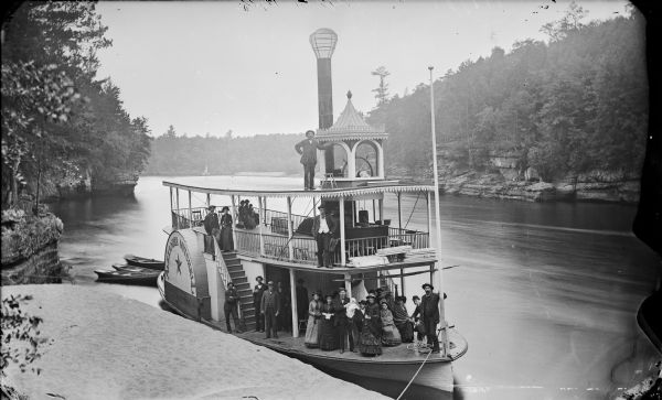 Elevated view from shoreline of the steamboat <i>Alexander Mitchell</i> docked at the edge of the river. Captain Dave and passengers are posing on board. Behind the stern are 3 empty row boats. In the background are bluffs and trees along the shoreline.