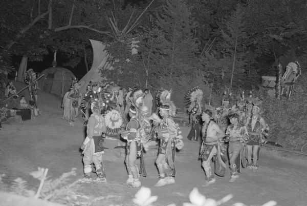 Slightly elevated view of Ho Chunk performing at the Stand Rock Indian Ceremonial at night. A group of men and a group of women dance in two circles, wearing Native American ceremonial clothing. On the right a man is playing a drum. On the left are a Ho Chunk man, women and child watching. In the background are trees, a tipi and bark house.<p>During its 78-year run (1919 to 1997) the Stand Rock Indian Ceremonial provided viewers a small glimpse into the ceremonies and dances performed by the Winnebago, Sioux, Kiowa and southwestern Native American tribes.</p>
