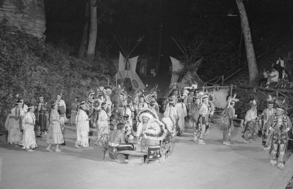 Slightly elevated view of Ho Chunk performing at the Stand Rock Indian Ceremonial at night. A group of men and women are dancing in a pattern, wearing Native American ceremonial clothing. In the foreground five men are drumming. On the right sitting on bleachers are spectators. In the background are two tipis, trees, a rock formation and spectator seating. Behind the tipis is a building.<p>During its 78-year run (1919 to 1997) the Stand Rock Indian Ceremonial provided viewers a small glimpse into the ceremonies and dances performed by the Winnebago, Sioux, Kiowa and southwestern Native American tribes.</p>