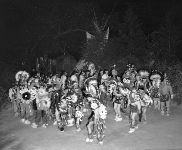 Slightly elevated view of Ho Chunk performing at the Stand Rock Indian Ceremonial at night. A group of men, women and children are dancing in a circle, wearing Native American ceremonial clothing. In the background is a tipi, trees, and bark house. On the left are some spectators and a Ho Chunk man.<p>During its 78-year run (1919 to 1997) the Stand Rock Indian Ceremonial provided viewers a small glimpse into the ceremonies and dances performed by the Winnebago, Sioux, Kiowa and southwestern Native American tribes.</p>