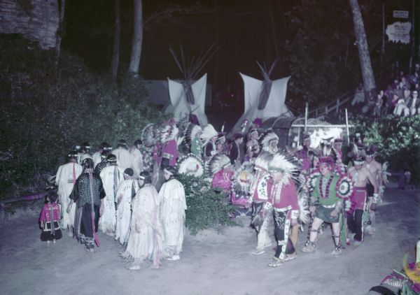 Slightly elevated view of the performance at the Stand Rock Indian Ceremonial. A group of men, women and children are performing the Green Corn Dance, moving in a circle and wearing Native American ceremonial clothing. The drummers are seated in the center of the circle among branches of foliage. In the background are two tipis, trees, and sandstone rock formations. Spectators are sitting on bleachers on the far right.<p>During its 78-year run (1919 to 1997) the Stand Rock Indian Ceremonial provided viewers a small glimpse into the ceremonies and dances performed by the Winnebago, Sioux, Kiowa and southwestern Native American tribes.</p>