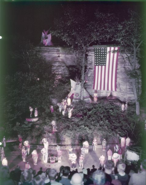 Elevated view of the closing performance at the Stand Rock Indian Ceremonial. A group of men, women and children are saluting the Flag of the United States, at night, wearing Native American ceremonial clothing. The flag is hanging in front of the sandstone rock formation, and a tipi is set up at the base. In the foreground are spectators sitting on bleachers.<p>During its 78-year run (1919 to 1997) the Stand Rock Indian Ceremonial provided viewers a small glimpse into the ceremonies and dances performed by the Winnebago, Sioux, Kiowa and southwestern Native American tribes.</p>