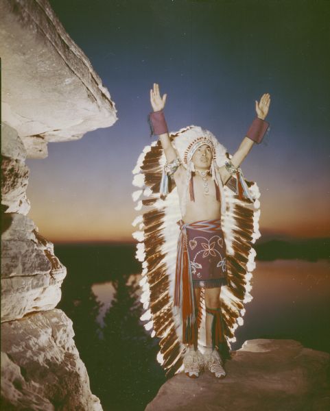 Sam (Carley) Blowsnake performing at the Stand Rock Indian Ceremonial, the Sunrise Call of the Zuni. He is dressed in Native American ceremonial clothing with a full-length feather headdress. He is standing on a rock outcropping with his back to the Wisconsin River which is far below. He is raising his arms over his head. On the left is a sandstone rock formation that is lit with artificial light.<p>During its 78-year run (1919 to 1997) the Stand Rock Indian Ceremonial provided viewers a small glimpse into the ceremonies and dances performed by the Winnebago, Sioux, Kiowa and southwestern Native American tribes.</p>