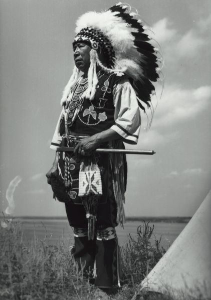 Chief Albert Yellow Thunder, Sr., standing in semi-profile, holding a peace pipe and wearing Native American ceremonial clothing. In the right foreground is the bottom edge of a tipi.