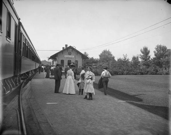View down platform, with railroad cars on the left, towards the Kilbourn railroad station. A crowd is standing along the platform. On the right is a lawn and trees.