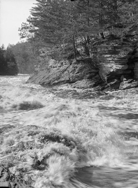 One side of stereograph of the Wisconsin River. View across rushing water towards a rock formation with trees on the opposite shoreline.