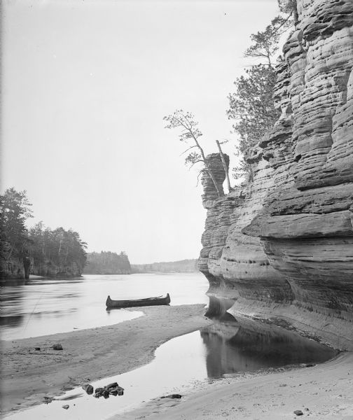Chimney Rock, south face. A canoe is beached on a sand bar near the foot. The river, more rock formations and trees are in the background.