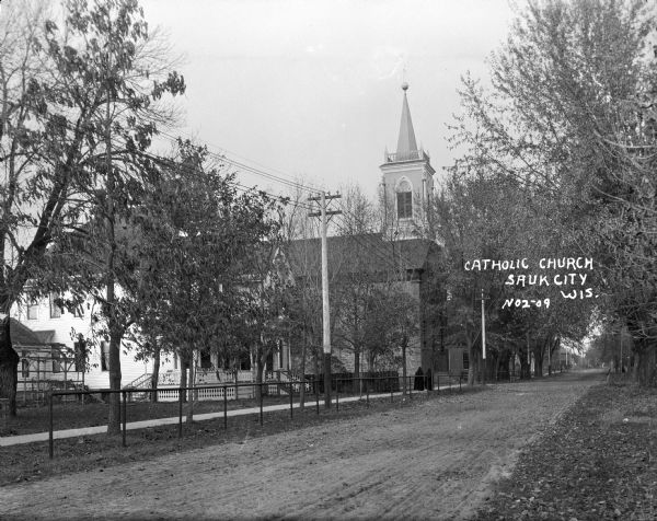 View down unpaved street towards a church. Two people, perhaps nuns, are walking on a sidewalk lined with metal railings. Caption reads: "Catholic Church, Sauk City, No2-09." On the far left is a house with a porch.