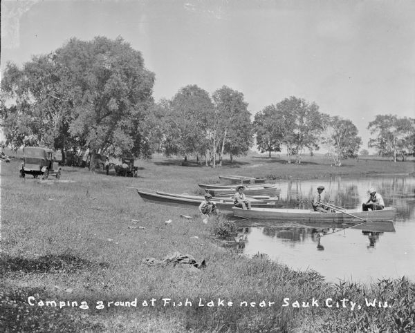 View along shoreline towards three boys sitting in a rowboat, and another boy sitting on the shore. A number of empty rowboats are pulled up on the shore behind them. On the left, four automobiles are parked on the grass, three of them in the shade of large trees, and three men are standing near the back of one of them. Caption reads: "Camping ground at Fish Lake near Sauk City, Wis." 