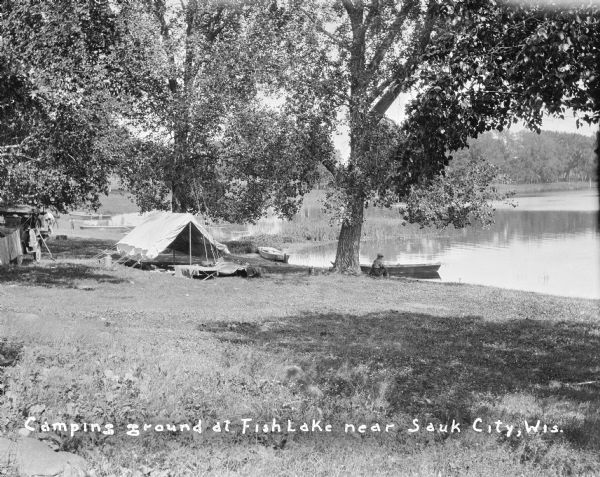 View down slope towards a man sitting on the shore of a lake beneath a tree, near a rowboat. Another boat is pulled up on the shore nearby. In the background on the left are more boats, men, children, automobiles, a large tent with cots in front, and laundry hanging on a line. Caption reads: "Camping ground at Fish Lake near Sauk City, Wis."