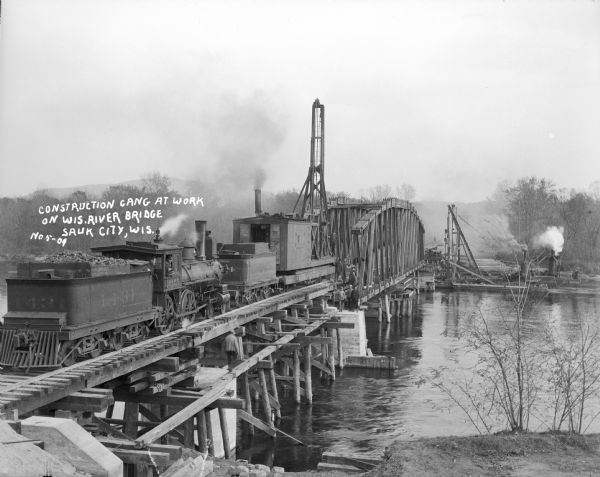 Elevated view of a work train on railroad tracks on a railroad bridge, over a river, that is under construction. Several workmen are standing and posing on timbers set up alongside the tracks. On the far shoreline smoke is rising from several steam powered machines. Caption reads: "Construction gang at work on Wis. River Bridge, Sauk City, Wis. No5-09."