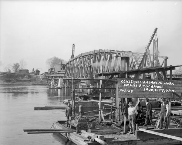 Elevated view of several workmen standing on a platform near the shoreline of the Wisconsin River during construction of a railroad bridge. There is a work train on the other end of the railroad bridge that appears to have a crane in the front. Caption reads: "Construction gang at work on Wis. River Bridge, Sauk City, Wis. No6-09."