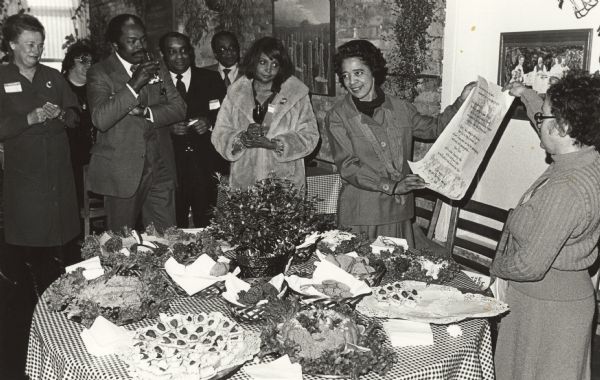 Liesl Blockstein of the Wisconsin Women's Political Caucus presenting guest-of honor, Vel Phillips, with an honorary poem at a reception at the L'Etoile Restaurant. In the foreground is a table holding a selection of hors' d'oeuvres and a flower arrangement. Vel is second from the right, holding one side of the poem.