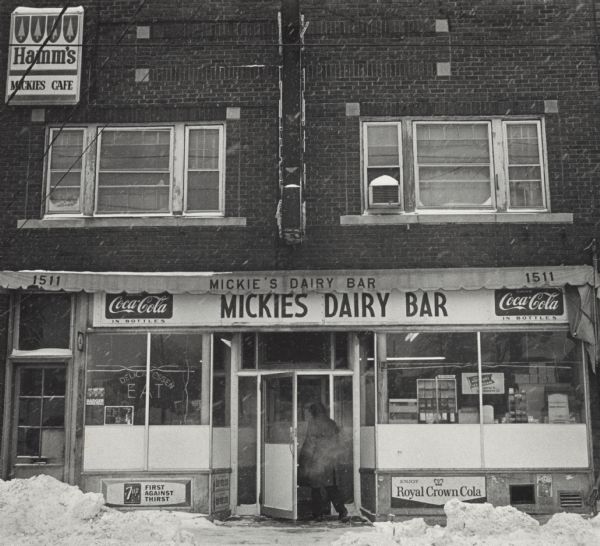 View across street towards a person entering Mickie's Dairy Bar in winter. The restaurant facade has signs advertising Coca-Cola, 7-Up, Royal Crown cola, and Hamms Beer.